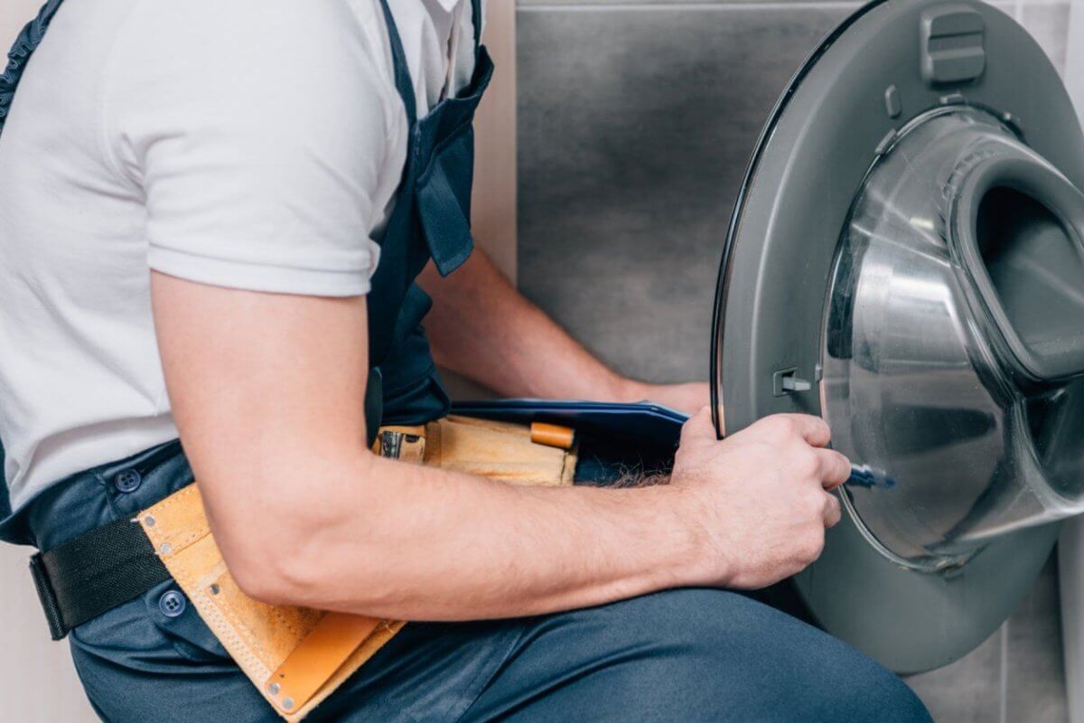 A person repairing a front-loading washing machine in Metro Manila. With a yellow tool belt brimming with various tools around their waist, they are closely examining the open door of the washing machine, appearing focused on providing top-notch Washing Machine Repair Services.
