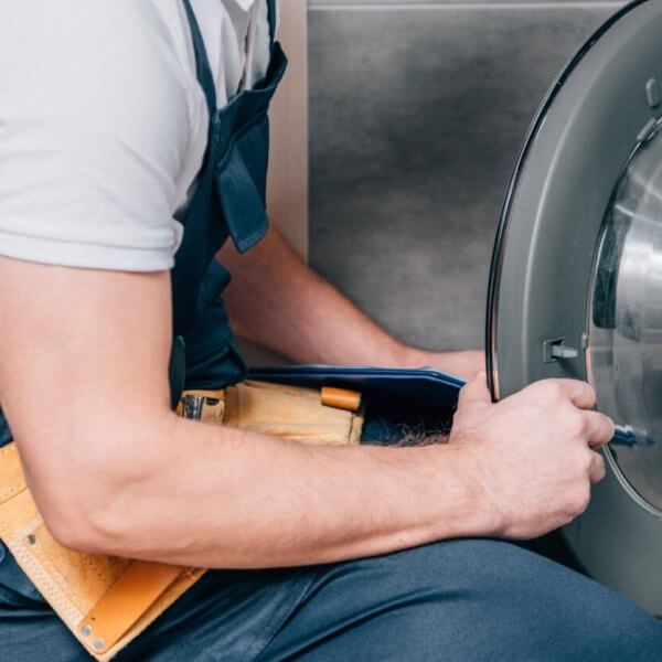 A person repairing a front-loading washing machine in Metro Manila. With a yellow tool belt brimming with various tools around their waist, they are closely examining the open door of the washing machine, appearing focused on providing top-notch Washing Machine Repair Services.