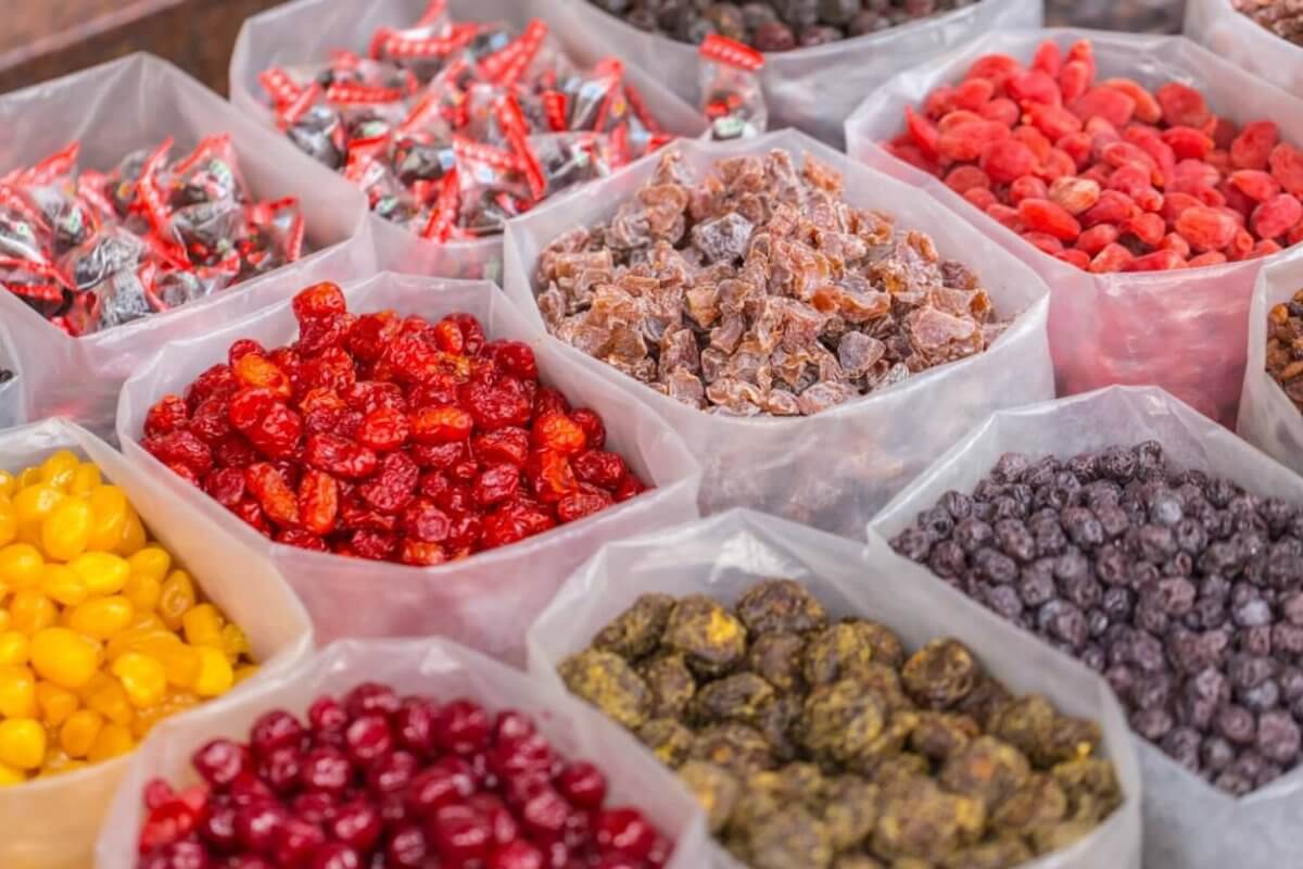 A market display of various dried fruits and candies from the best wholesale food suppliers in Metro Manila. Clear plastic bins are filled with colorful selections such as red cherries, yellow berries, purple raisins, and multicolored preserved fruits. Wrapped candies are also visible in the background, creating a vibrant and inviting scene.