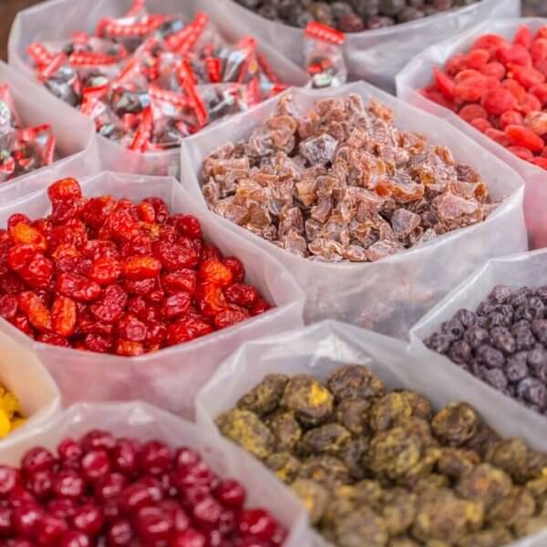 A market display of various dried fruits and candies from the best wholesale food suppliers in Metro Manila. Clear plastic bins are filled with colorful selections such as red cherries, yellow berries, purple raisins, and multicolored preserved fruits. Wrapped candies are also visible in the background, creating a vibrant and inviting scene.