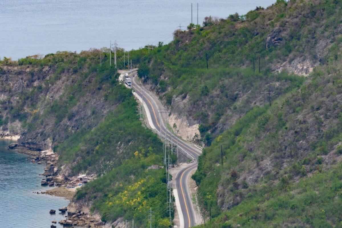 A winding road cuts through the lush, hilly terrain of Soccsksargen along the coastline. The road closely follows the contour of the land, with the sea visible on the left. Sparse greenery dots the slopes, and a few vehicles travel on this Region 12 route. The calm sea and green hills create a tranquil, scenic view.