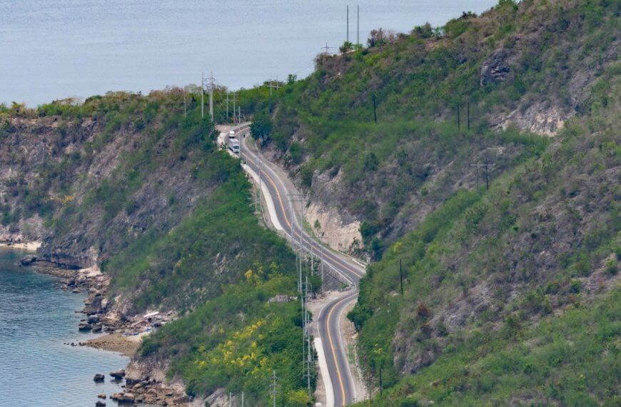 A winding road cuts through the lush, hilly terrain of Soccsksargen along the coastline. The road closely follows the contour of the land, with the sea visible on the left. Sparse greenery dots the slopes, and a few vehicles travel on this Region 12 route. The calm sea and green hills create a tranquil, scenic view.