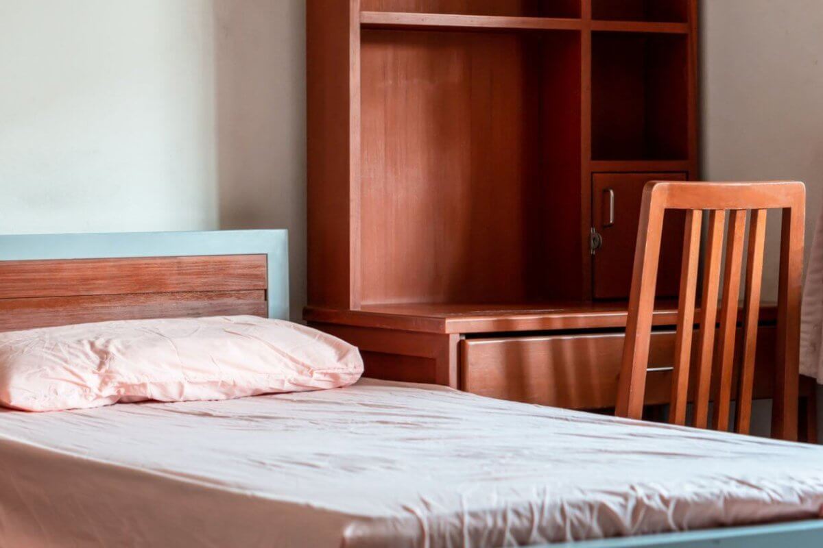 A neatly made single bed with a light pink pillow and blanket, positioned next to a wooden desk and chair set. The desk has storage compartments and a shelf unit above it. A curtain-draped window is partially visible on the right, letting in soft natural light—perfect for your stay in Metro Manila's best apartelle.