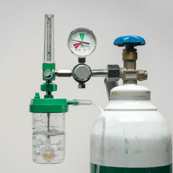 A medical oxygen tank with a pressure gauge, flow meter, and humidifier bottle attached. The tank is white with a green band near the top, and the attached components are mostly green and silver, with a blue knob. Commonly available from oxygen tank suppliers in Metro Manila, the backdrop is plain light gray.