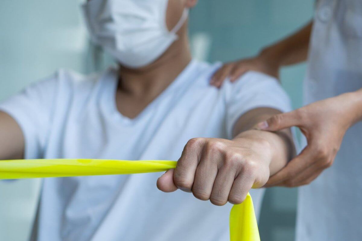 a person wearing a face mask is exercising with a yellow resistance band. Another individual, also masked, stands beside them with one hand on their back, providing support and guidance. The setting appears to be a healthcare or rehabilitation environment offering Physical Therapy Services.