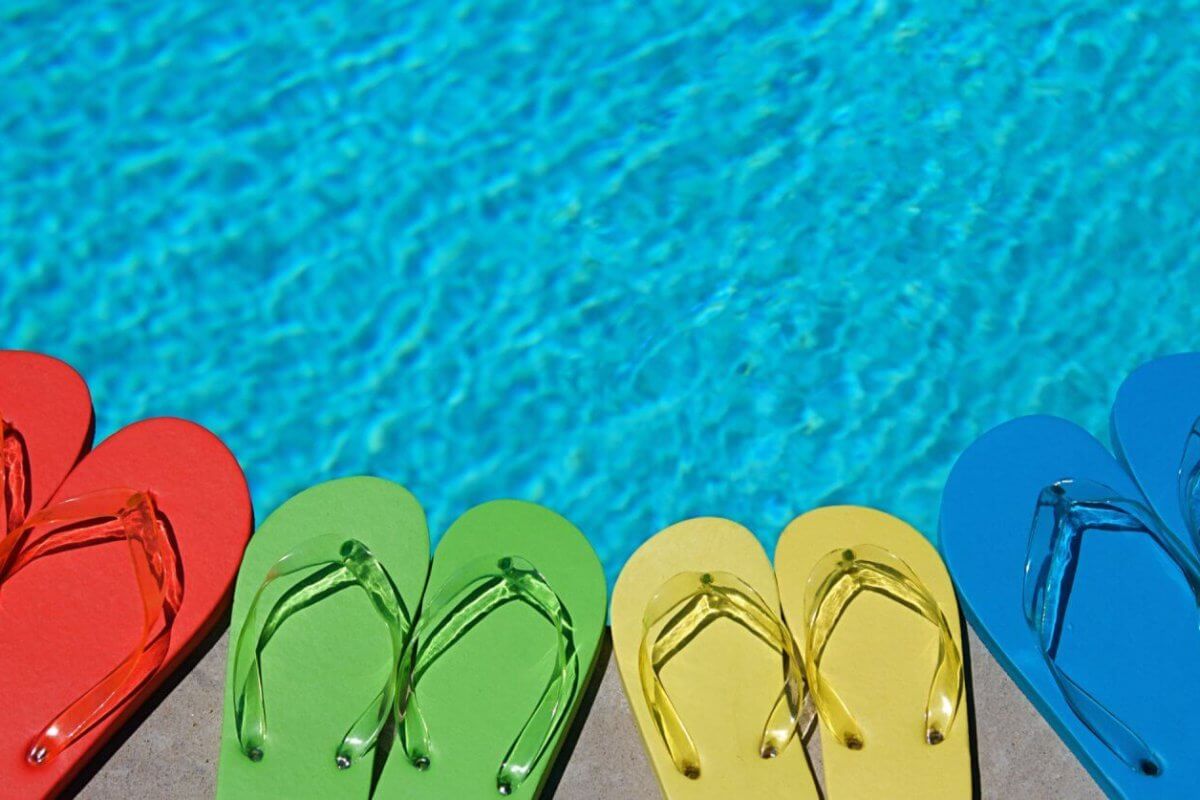Four pairs of flip-flops lined up near a swimming pool at one of the best resorts. The flip-flops are in different colors: red, green, yellow, and blue. The clear blue pool water reflects the sunlight, evoking a bright, summery day perfect for an unforgettable escape.