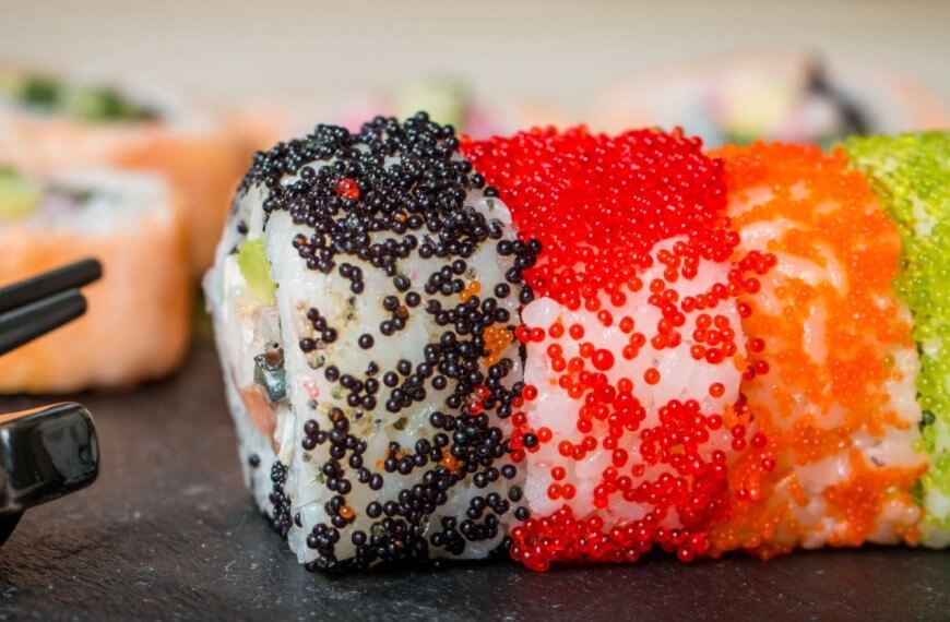 Close-up of sushi rolls from Mitsukoshi BGC decorated with black, red, and orange fish roe on a slate surface. The rolls have a visible mix of rice and seaweed, and various other ingredients inside, with out-of-focus sushi pieces seen in the background. Black chopsticks are placed to the left.