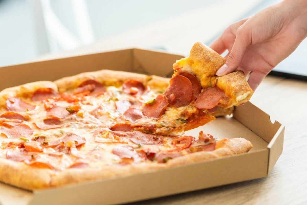 A hand is picking up a slice of pepperoni pizza from an open cardboard pizza box. The pizza has a golden-brown crust, melted cheese, and pepperoni slices. Enjoying this mouthwatering treat at one of the best restaurants in Cubao near Farmers Plaza adds to the delightful indoor setting.