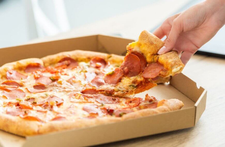 A hand is picking up a slice of pepperoni pizza from an open cardboard pizza box. The pizza has a golden-brown crust, melted cheese, and pepperoni slices. Enjoying this mouthwatering treat at one of the best restaurants in Cubao near Farmers Plaza adds to the delightful indoor setting.