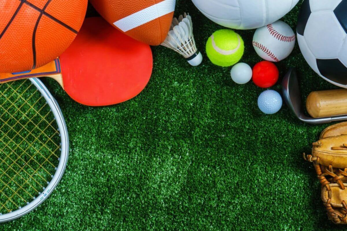 A variety of sports equipment arranged on a green grass surface. Items include a basketball, yoga mat, American football, soccer ball, badminton birdie, table tennis paddle, baseball glove, tennis ball, golf balls, cricket bat. Perfect for enthusiasts in Metro Manila's sports centers.