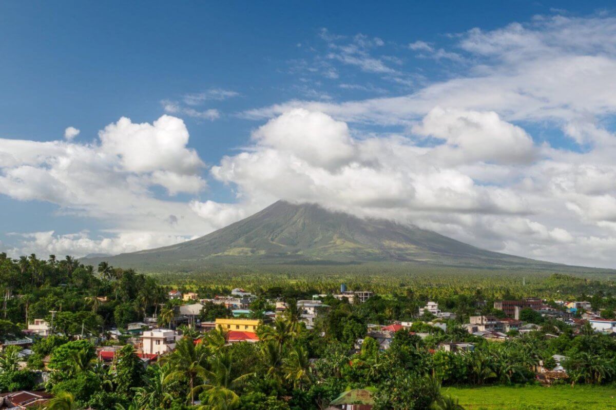 A picturesque landscape in the Bicol Region features a dormant volcano enveloped in clouds with a lush, green forest at its base. In the foreground, a small town with colorful buildings and rooftops. The sky is bright blue with scattered white clouds, creating a serene atmosphere.