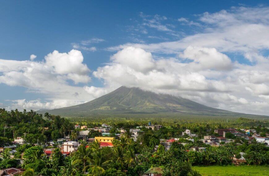 A picturesque landscape in the Bicol Region features a dormant volcano enveloped in clouds with a lush, green forest at its base. In the foreground, a small town with colorful buildings and rooftops. The sky is bright blue with scattered white clouds, creating a serene atmosphere.