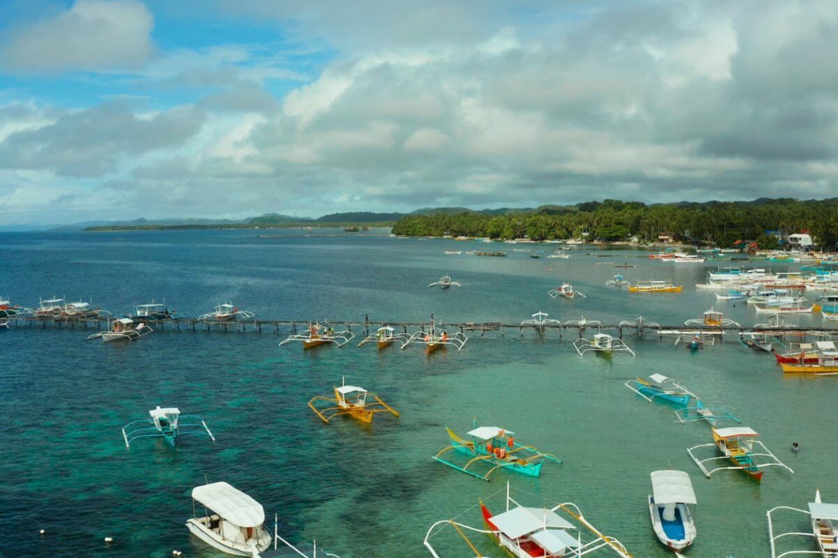 The image captures a coastal scene in the Caraga Region with numerous colorful outriggers and small boats docked or moored in clear, calm waters. The backdrop includes lush green trees, a cloudy blue sky, and distant landforms, creating a serene and picturesque maritime environment.