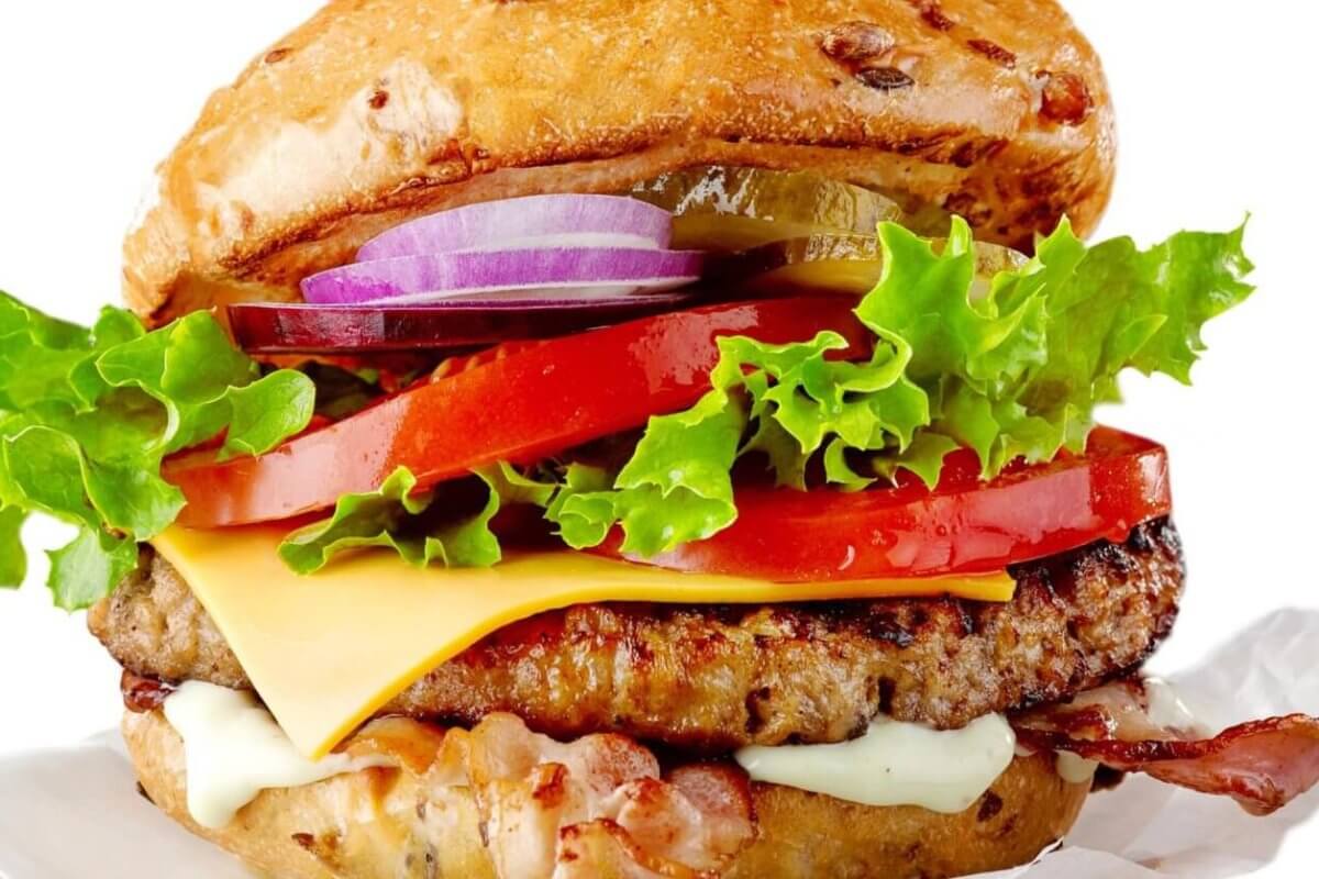 A colorful, juicy cheeseburger from one of the best restaurants in Imus Cavite is shown stacked high with layers of fresh, crisp lettuce, sliced red tomatoes, melted cheese, savory beef patty, crispy bacon, and sliced red onions. The bun is lightly toasted and speckled with sesame seeds. The burger is set against a white background.