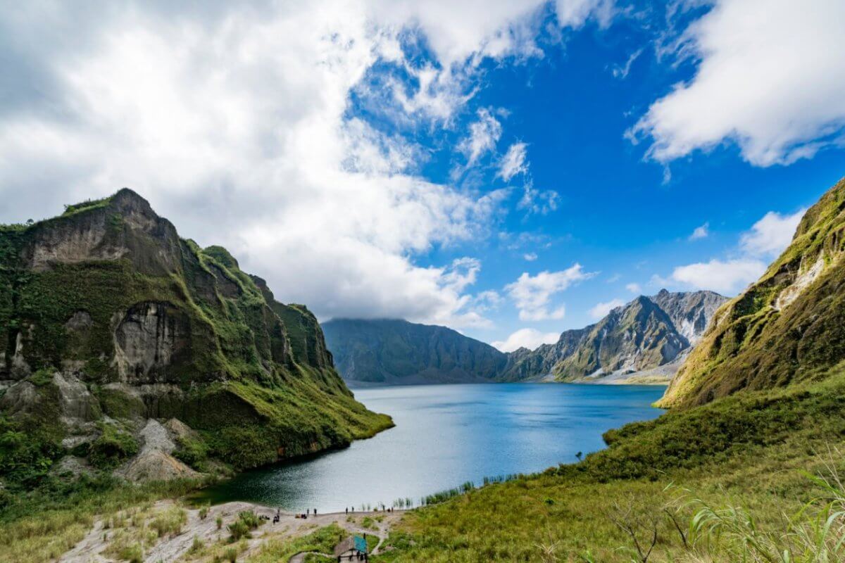 A pristine lake is nestled between towering, green-covered mountains in Region 3 under a partly cloudy sky. The vibrant blue water reflects the surrounding cliffs and the azure of the sky. Lush vegetation covers the mountainsides, and a few people are visible on a path near the lake.