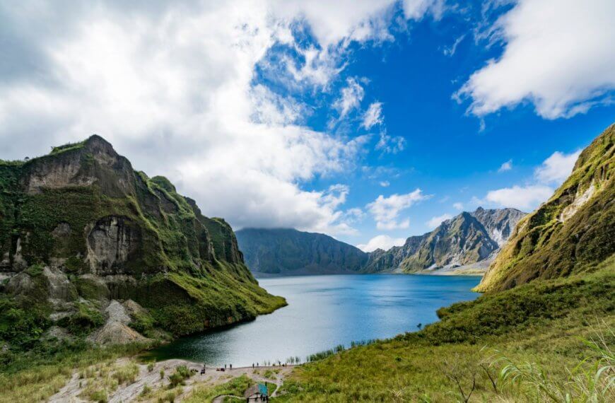 A pristine lake is nestled between towering, green-covered mountains in Region 3 under a partly cloudy sky. The vibrant blue water reflects the surrounding cliffs and the azure of the sky. Lush vegetation covers the mountainsides, and a few people are visible on a path near the lake.