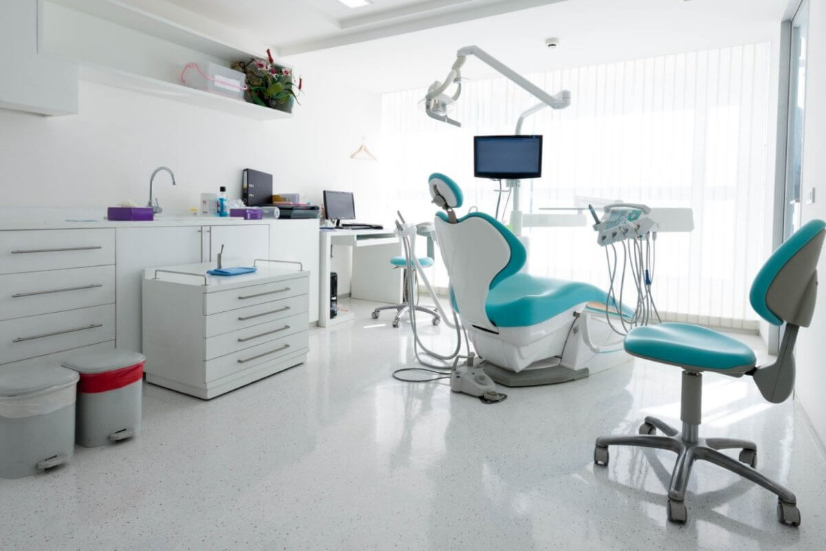 A modern dental office in Iligan City with white walls and floors. The room features a dental chair with an overhead light, a monitor, and various instruments. Two additional blue and white chairs are present. Cabinets line the left wall, and a desk with a computer is in the background — one of the best dental clinics around.