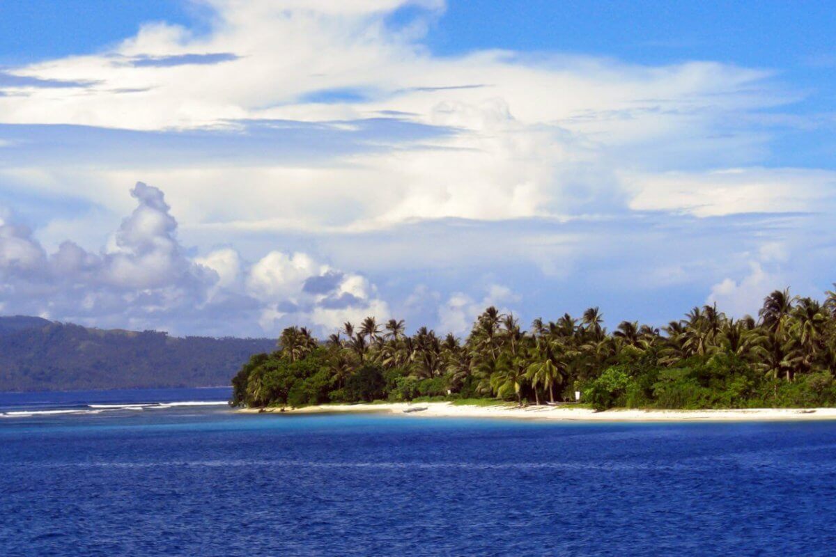 A picturesque tropical island in Eastern Visayas is surrounded by blue ocean water. The coastline, within Region 8's stunning locales, is lined with lush green palm trees and greenery that stands out against the sandy beach. In the background, hills and a partly cloudy sky with scattered white clouds meet the horizon.