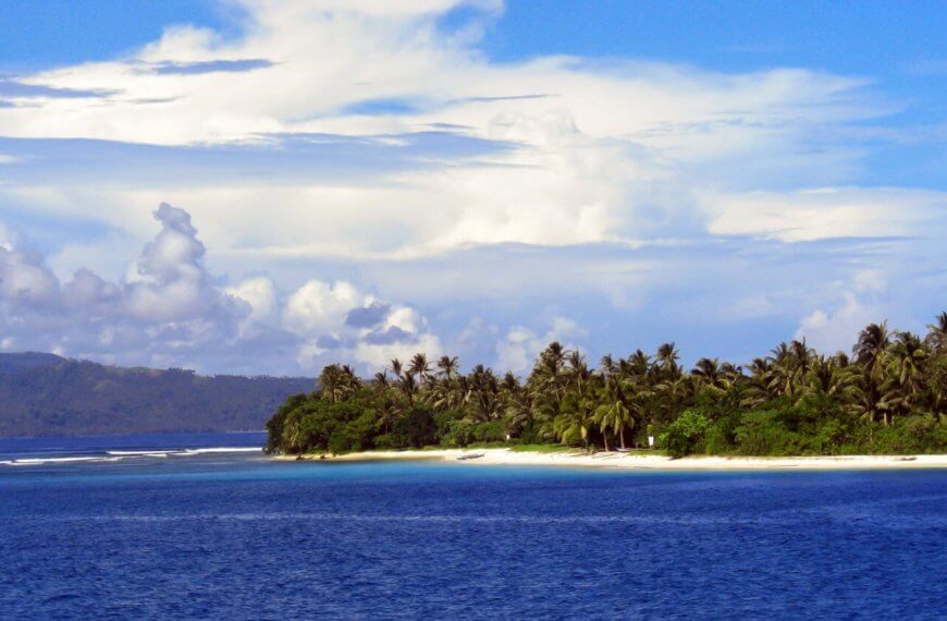 A picturesque tropical island in Eastern Visayas is surrounded by blue ocean water. The coastline, within Region 8's stunning locales, is lined with lush green palm trees and greenery that stands out against the sandy beach. In the background, hills and a partly cloudy sky with scattered white clouds meet the horizon.