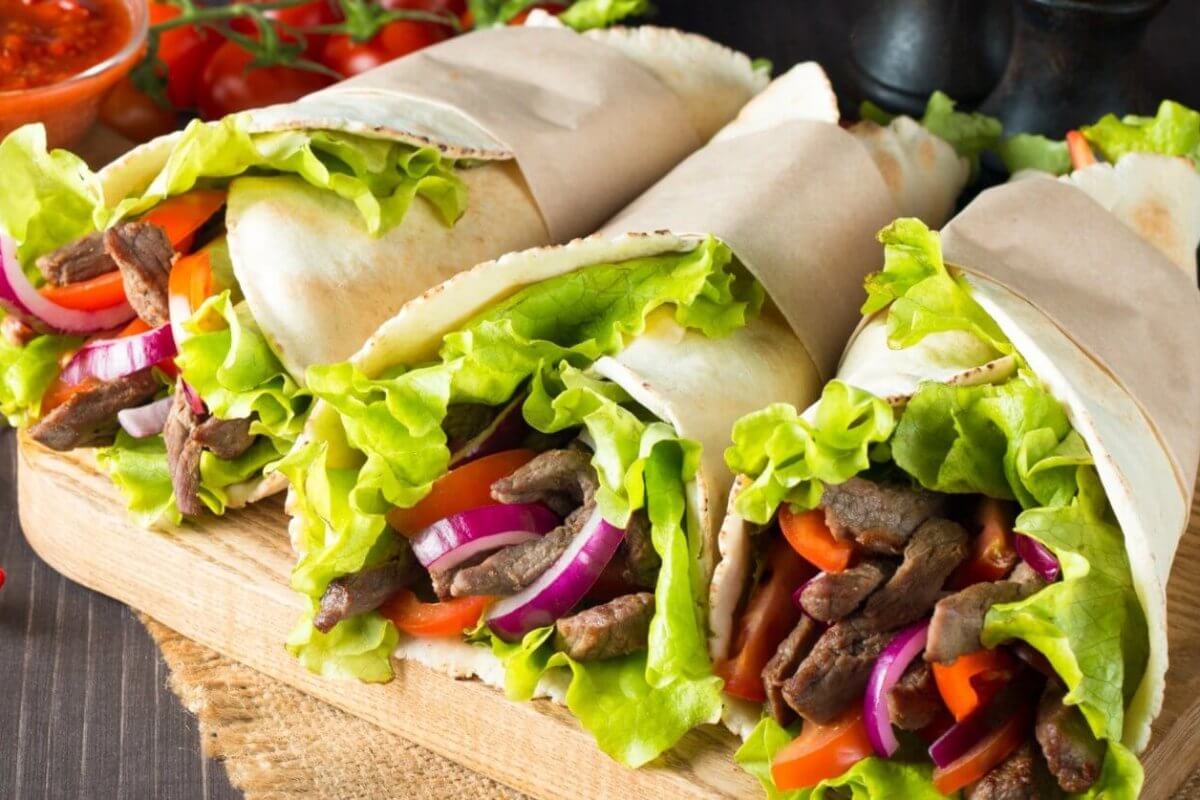 Three wraps filled with seasoned beef strips, fresh lettuce, sliced red onions, and red bell peppers are displayed on a wooden board at one of Makati's renowned Greenbelt Mall restaurants. The wraps are partially wrapped in parchment paper. In the background, there is a bowl of red sauce, cherry tomatoes, and a whole red chili pepper.