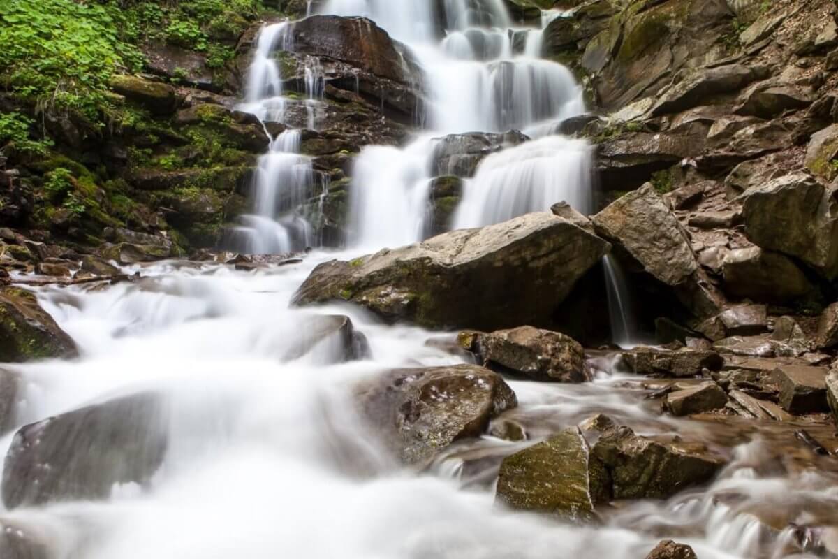 A cascading waterfall flows over multiple rocky tiers surrounded by lush green foliage, a highlight among Iligan's tourist attractions. The water appears smooth and silky, contrasting with the rugged, moss-covered rocks. The scene conveys a tranquil and refreshing natural environment.