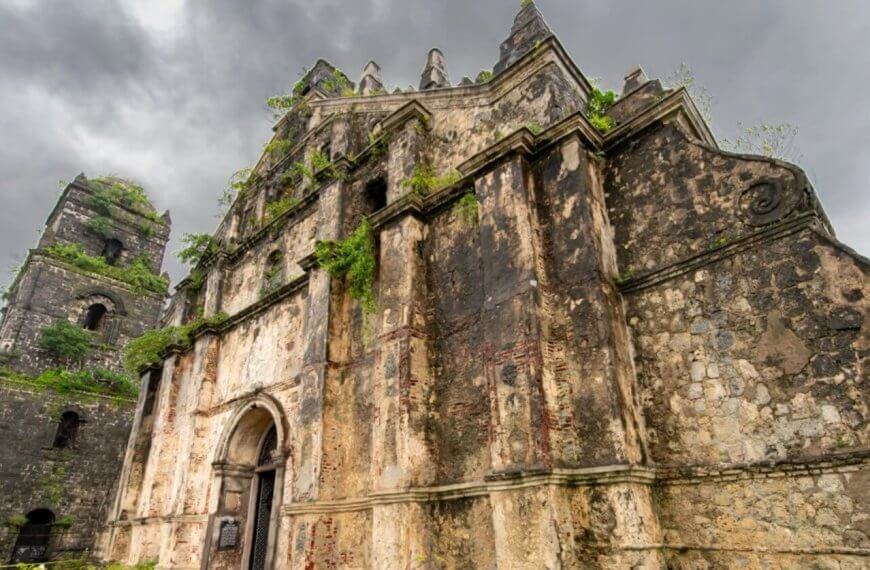 An ancient, weathered church facade stands against a dark, cloudy sky in the Ilocos Region. The stone structure features arches, ornate carvings, and an adjacent bell tower. Vines and plants grow out of the stone, emphasizing its aged and abandoned appearance. The scene exudes a historical and dramatic atmosphere.