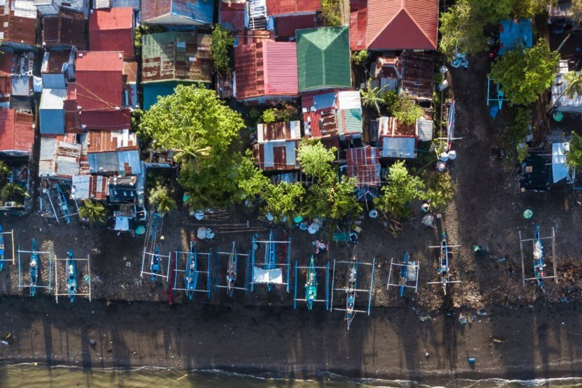Aerial view of a coastal village in Region 4B, featuring numerous small, colorful houses with tin roofs. Many houses are surrounded by trees. Fishing boats line the dark, sandy shoreline, casting long shadows. The beach rests in the foreground, transitioning into the bluish water at the bottom of the image.