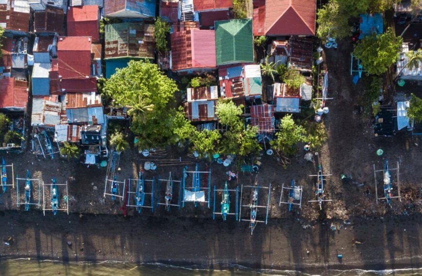 Aerial view of a coastal village in Region 4B, featuring numerous small, colorful houses with tin roofs. Many houses are surrounded by trees. Fishing boats line the dark, sandy shoreline, casting long shadows. The beach rests in the foreground, transitioning into the bluish water at the bottom of the image.