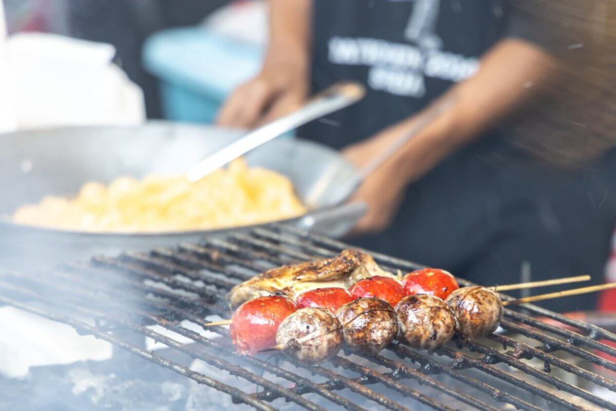 A person grills skewers of mushrooms and cherry tomatoes over a flame. In the background, another person is cooking a dish in a large wok. Both individuals are partially out of focus, with the grilling food as the main subject. The lively scene seems to capture one of Quezon City's best outdoor market spots.

