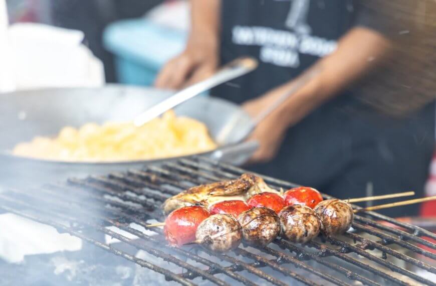 A person grills skewers of mushrooms and cherry tomatoes over a flame. In the background, another person is cooking a dish in a large wok. Both individuals are partially out of focus, with the grilling food as the main subject. The lively scene seems to capture one of Quezon City's best outdoor market spots.