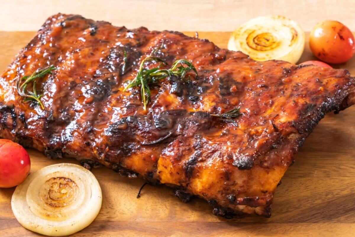 A slab of grilled barbecue ribs rests on a wooden cutting board, adorned with char marks and a coating of sauce. Garnishes of roasted cherry tomatoes, whole onion slices, and rosemary sprigs are arranged around the ribs, enhancing the visual appeal of the dish—perfect for dining at SM Fairview restaurants in Quezon City.
