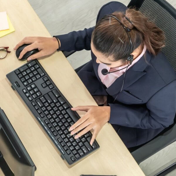 A person wearing a headset is seated at a desk using a computer keyboard and mouse, possibly working as one of the best virtual assistants. The individual is dressed in business attire. The desk has eyeglasses and sticky notes on it. The scene is viewed from an overhead perspective.