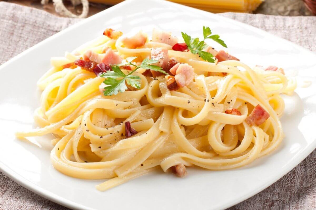 A plate of pasta carbonara on a white square dish from one of Bridgetowne's charming restaurants. The dish features fettuccine noodles coated in a creamy sauce, sprinkled with bacon pieces and black pepper, and garnished with fresh parsley. Set on a brown cloth with a rustic wooden background.