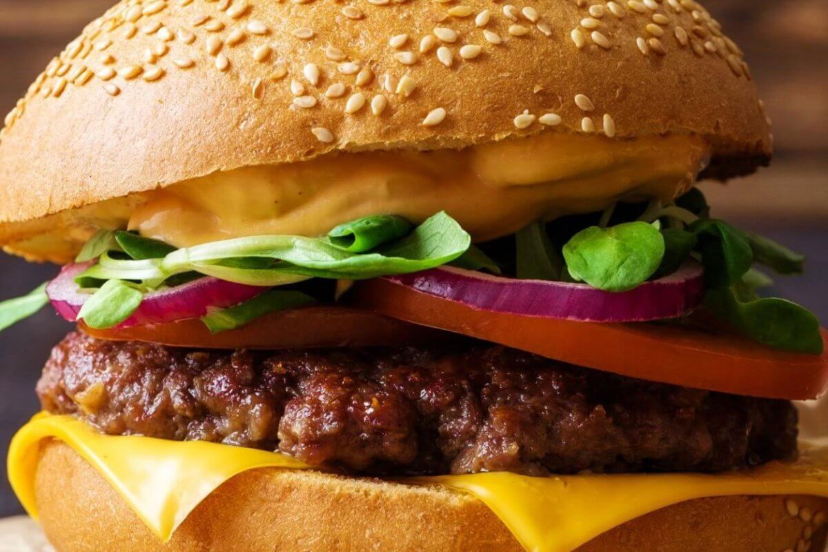 A close-up of one of Metro Manila's best burgers: a sesame seed bun enveloping layers of melted cheese, a juicy beef patty, sliced red onions, a tomato slice, fresh spinach leaves, and a generous layer of creamy sauce. The slightly blurred background accentuates the burger's vibrant textures and colors.