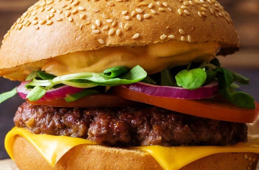 A close-up of one of Metro Manila's best burgers: a sesame seed bun enveloping layers of melted cheese, a juicy beef patty, sliced red onions, a tomato slice, fresh spinach leaves, and a generous layer of creamy sauce. The slightly blurred background accentuates the burger's vibrant textures and colors.