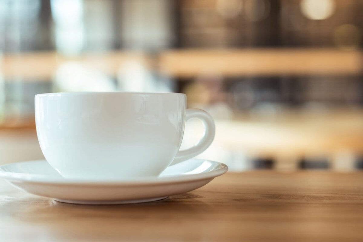 A white coffee cup rests on a matching saucer on a wooden table, embodying the essence of Metro Manila's best coffee. The blurred background, with warm tones and hints of light, suggests a cozy cafe atmosphere, evoking a sense of calm and relaxation.