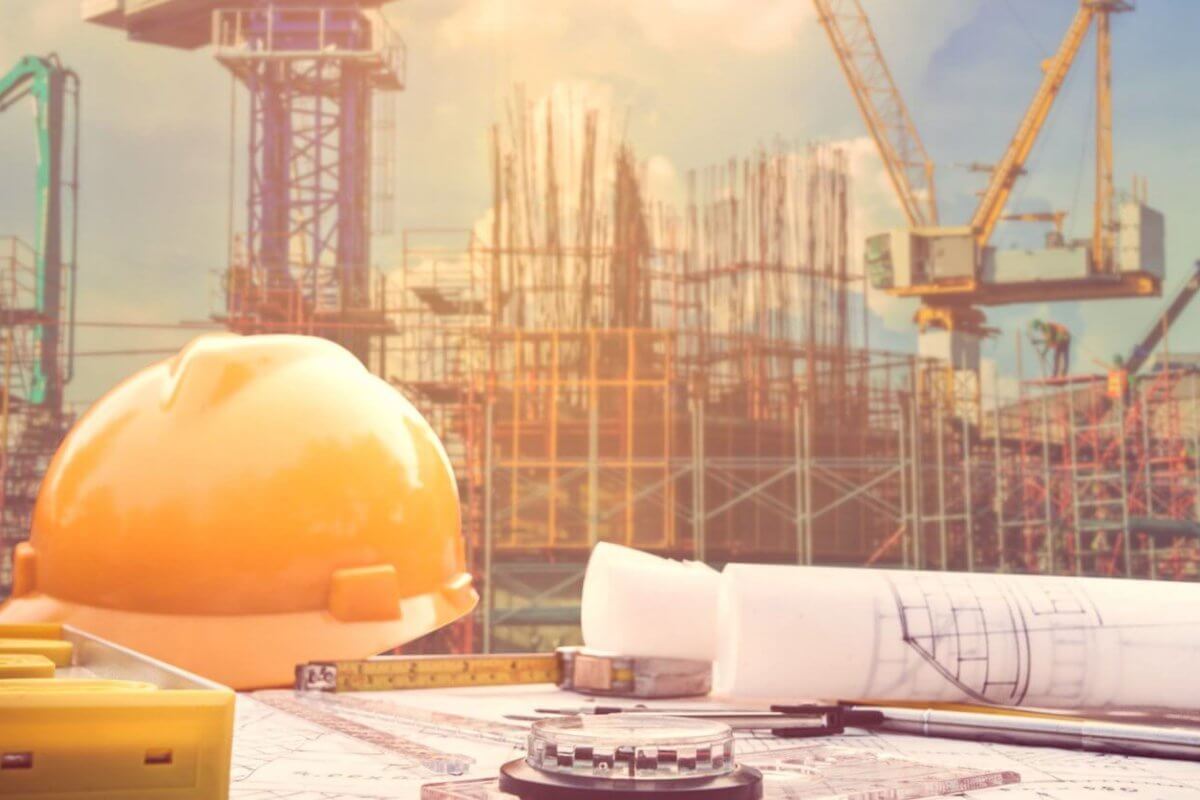A construction site with cranes and scaffolding rises under Manila's blue sky. In the foreground, a yellow hard hat, rolled blueprints, a measuring tape, and a compass rest on a table, symbolizing architectural planning backed by construction supply companies known for quality and reliability.
