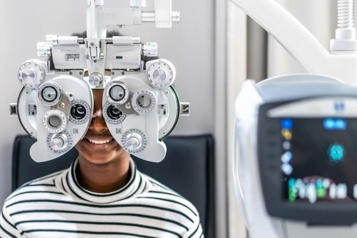 In one of Metro Manila's best eye clinics, a person in a striped shirt sits behind a phoropter, undergoing an eye examination. The focus is on the device with its various dials and lenses, while a blurred vision-testing machine looms in the foreground. The scene is set in a bright, clinical room.