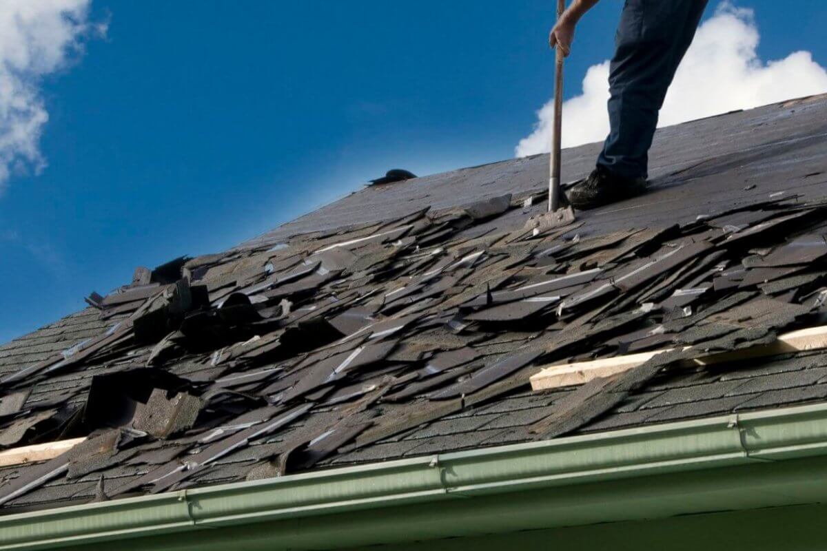 A person is removing old shingles from a sloped roof with a tool sourced from premium roofing suppliers. The roof, with its damaged and scattered shingles, needs attention. Wearing jeans, the person stands against a clear blue sky with some clouds, while a green gutter lines the edge of the roof.