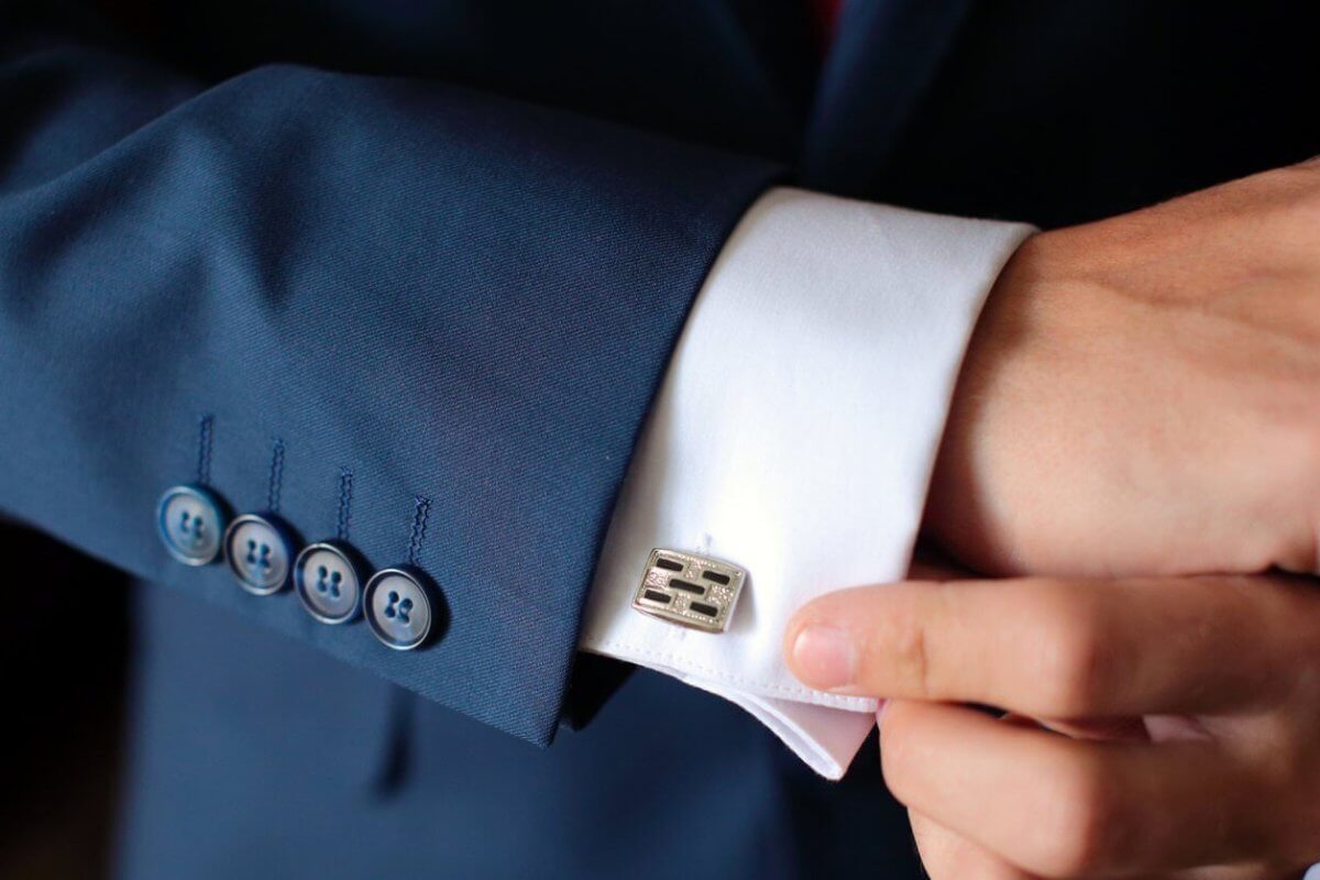 A person expertly adjusts a metallic, rectangular cufflink on a fitted white dress shirt. The tailored sleeve, part of a dark blue suit with four blue buttons, exudes elegance. The setting is shadowed and elusive, reminiscent of Metro Manila's sophisticated style.