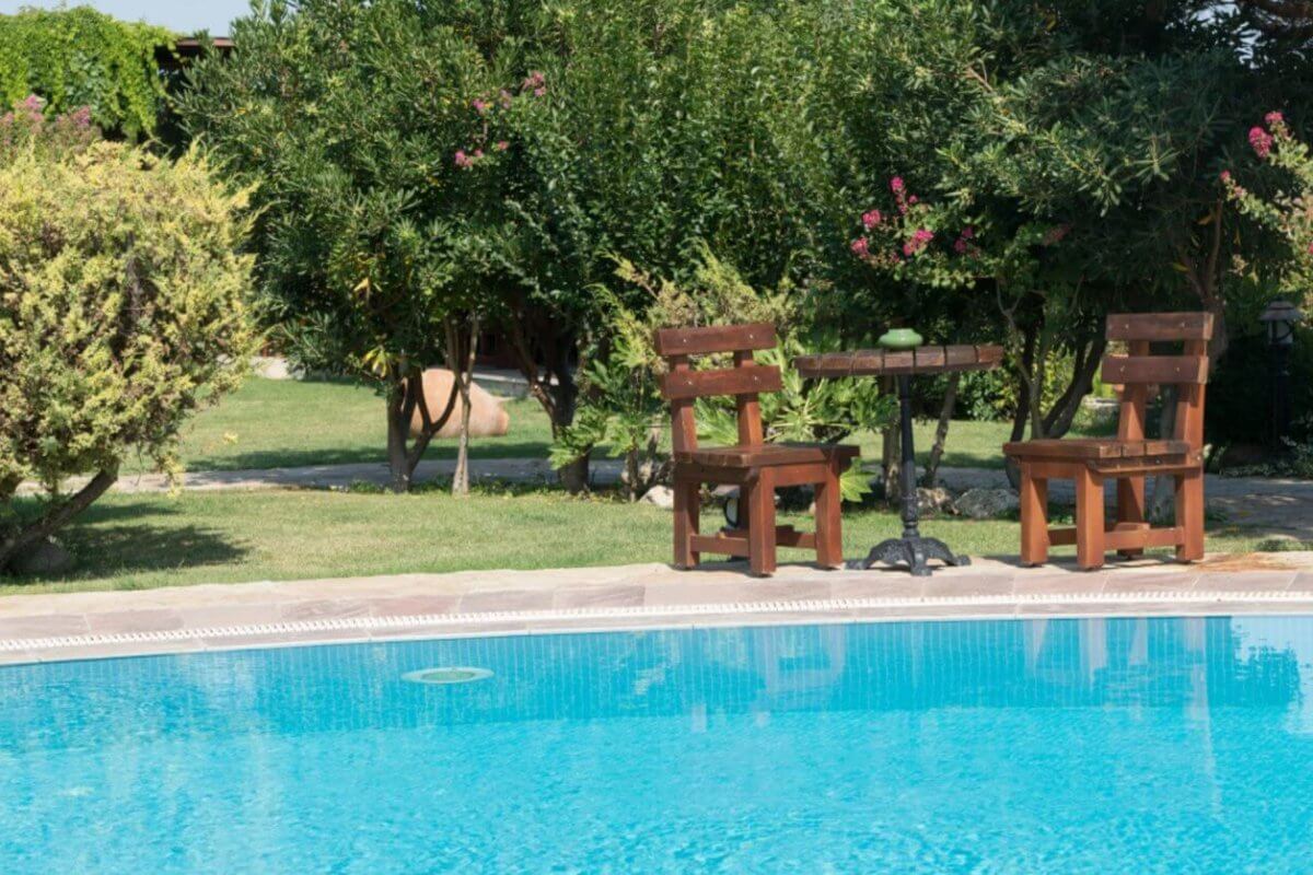 A serene outdoor scene with a clear blue swimming pool in the foreground, reminiscent of Calamba's famous hot springs. In the background, two wooden chairs and a small round table rest on a lush lawn in Laguna. Surrounding bushes and trees add nature's touch and privacy to the setting.