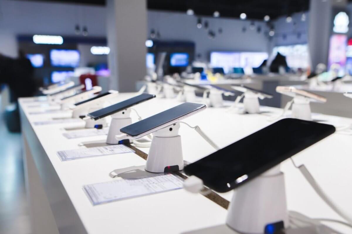 Smartphones are displayed on a white counter in a DITO Experience Store, each mounted on a stand with security features. The store, located in the Philippines, has bright lighting and a blurred background showcasing more electronic devices, with price tags accompanying each phone.