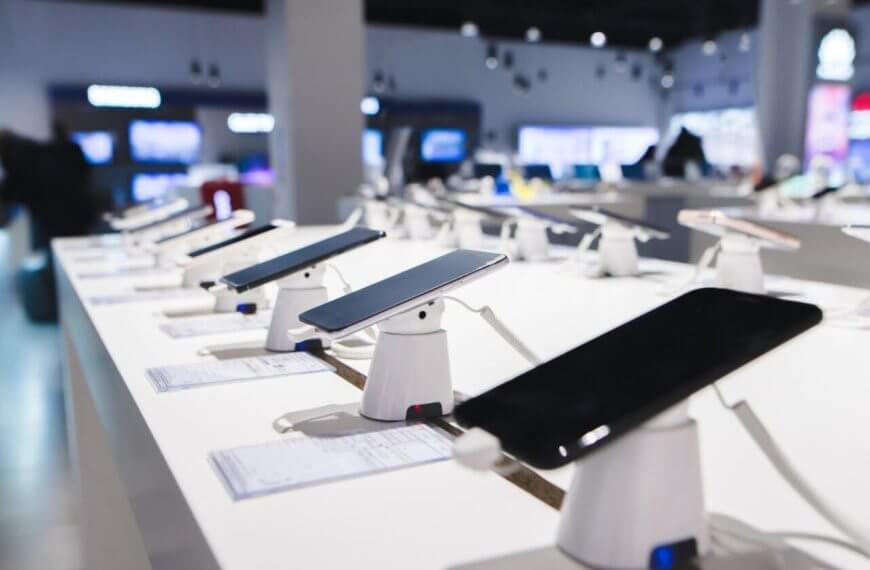 Smartphones are displayed on a white counter in a DITO Experience Store, each mounted on a stand with security features. The store, located in the Philippines, has bright lighting and a blurred background showcasing more electronic devices, with price tags accompanying each phone.