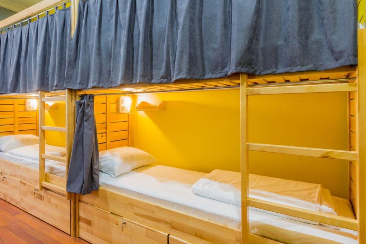 Cozy bunk beds with wooden frames, each adorned with navy curtains, pillows, and lamps, rest against a yellow wall in the best dormitory in Metro Manila.