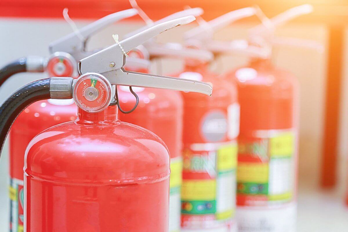Close-up of red fire extinguishers, supplied by top Fire Extinguisher Suppliers in Metro Manila, standing in a row. The foreground extinguisher is in focus, its nozzle and pressure gauge clear, while others blur into the background. The scene is brightly lit with a warm glow in the top right corner.
