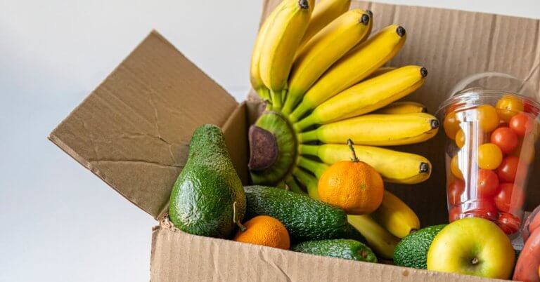 A cardboard box from a food distributor in Metro Manila is filled with bananas, avocados, and oranges. A plastic cup containing red and yellow cherry tomatoes is also visible. An apple rests at the front of the open box, showcasing the colorful and fresh produce against a neutral background.