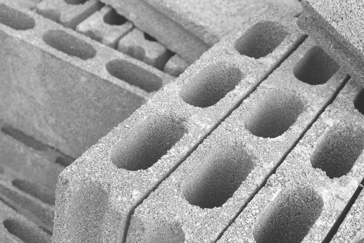 A close-up of stacked concrete cinder blocks, sourced from leading hollow block suppliers in Metro Manila, forms a monochrome setting. These blocks, with three elongated oval holes and rough textures, are arranged diagonally. Some are fully visible while others are partially obscured, crafting a repetitive geometric pattern.