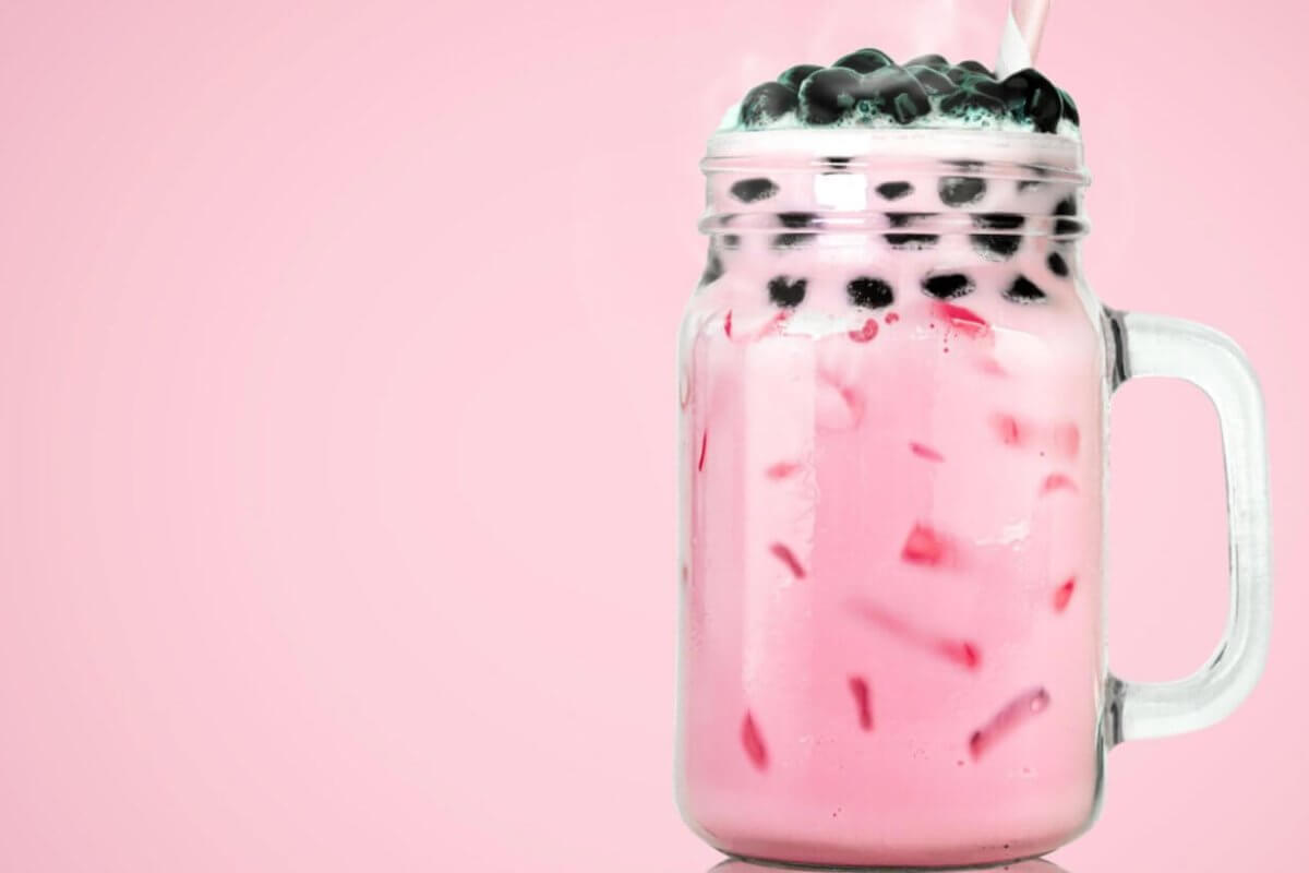 A clear glass mason jar filled with pink bubble tea from top Milktea suppliers, featuring black tapioca pearls on top. The drink contains ice cubes and is set against a solid pink background. A straw is inserted into the jar, evoking the refreshing, sweet flavors cherished in Metro Manila.