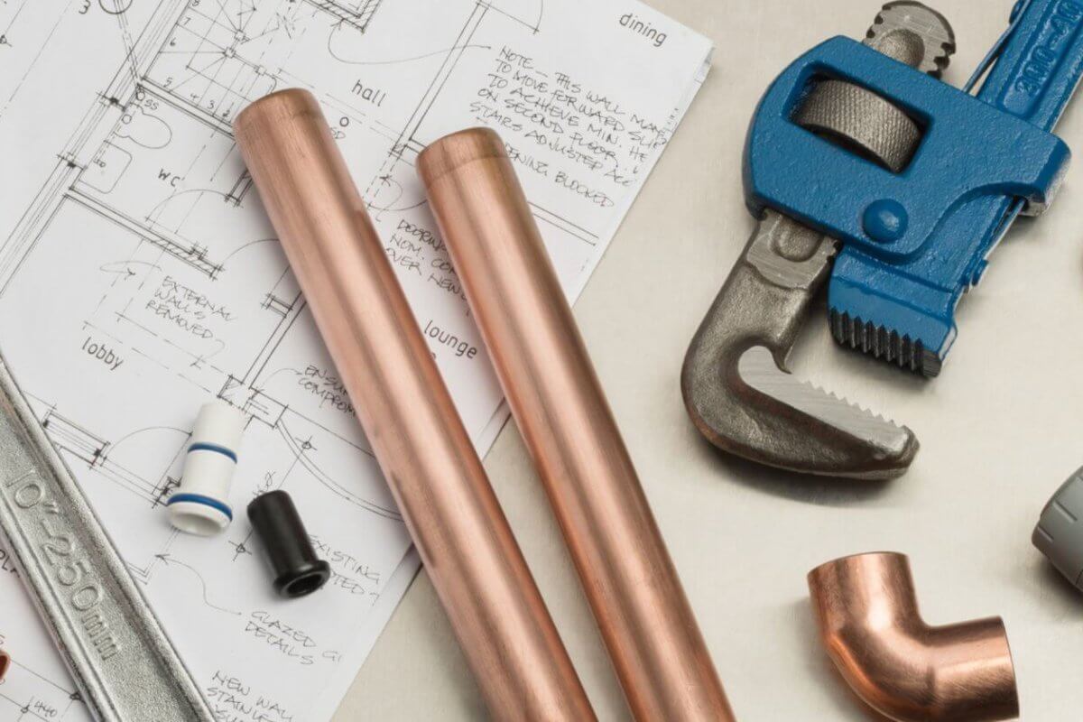 Plumbing tools and materials, ideal for plumbing services in Metro Manila, are neatly arranged on an architectural blueprint. Items include a large adjustable wrench, two copper pipes, a blue pipe wrench, copper fittings, and various pipe components set against detailed floor plans and elevations.