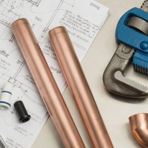 Plumbing tools and materials, ideal for plumbing services in Metro Manila, are neatly arranged on an architectural blueprint. Items include a large adjustable wrench, two copper pipes, a blue pipe wrench, copper fittings, and various pipe components set against detailed floor plans and elevations.